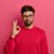 No problem concept. Bearded man makes okay gesture, has everything under control, all fine gesture, wears spectacles and jumper, poses against pink background, says I got this, guarantees something